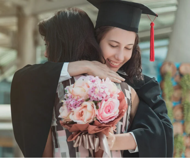 Female in graduation cap and gown, holding flowers, hugging another female