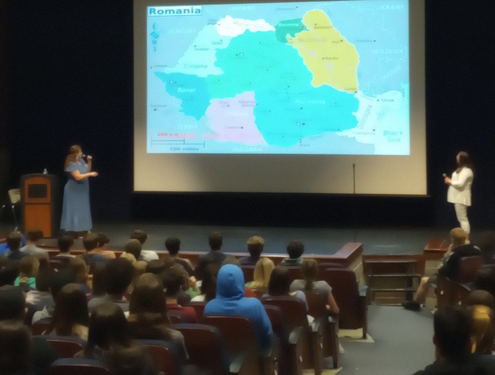 Two women standing in front of a large screen with a map of the Europe on it the screen on it, while people are sitting in the audience watching. 