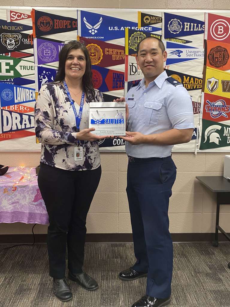A women and a man holding  a certificate with college pennants in the background. The man is wearing an Air Force Uniform